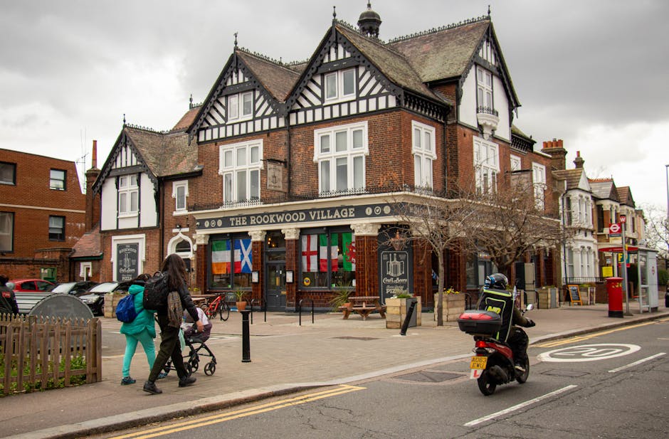 A multi-story brick building with a Tudor-style architectural design featuring black timber framing and white plaster infill, situated on a street corner in an urban area. The ground floor hosts a pub or restaurant named 'The Rookwood Village,' with a black sign and gold lettering, and multiple national flags including the Irish, Scottish, and Welsh flags hanging beneath the window. In front of the building, there is a paved sidewalk with wooden picnic tables and black bollards, and a leafless tree adjacent to the entrance. A couple of pedestrians, including a woman pushing a stroller and a person wearing a teal jacket, are walking along the sidewalk. On the street, a motorcyclist wearing a black helmet and jacket is approaching, while parked cars are visible in the background. The environment appears overcast with cloudy skies, creating a muted lighting atmosphere. The scene reflects a typical urban setting suitable for private waste handling or independent rubbish collection, with no visible litter or waste, emphasizing the clean, managed streetscape associated with professional rubbish removal services occasionally operating in such areas.