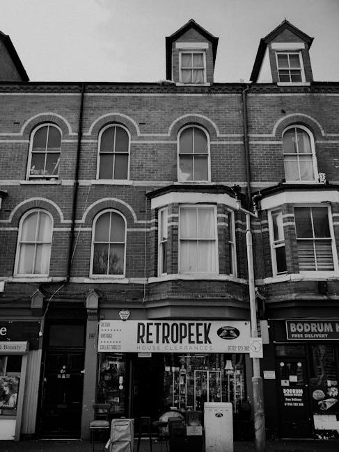 A multi-story brick building with a Tudor-style architectural design featuring black timber framing and white plaster infill, situated on a street corner in an urban area. The ground floor hosts a pub or restaurant named 'The Rookwood Village,' with a black sign and gold lettering, and multiple national flags including the Irish, Scottish, and Welsh flags hanging beneath the window. In front of the building, there is a paved sidewalk with wooden picnic tables and black bollards, and a leafless tree adjacent to the entrance. A couple of pedestrians, including a woman pushing a stroller and a person wearing a teal jacket, are walking along the sidewalk. On the street, a motorcyclist wearing a black helmet and jacket is approaching, while parked cars are visible in the background. The environment appears overcast with cloudy skies, creating a muted lighting atmosphere. The scene reflects a typical urban setting suitable for private waste handling or independent rubbish collection, with no visible litter or waste, emphasizing the clean, managed streetscape associated with professional rubbish removal services occasionally operating in such areas.