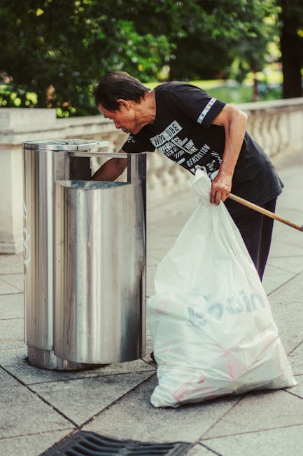 A man cleaning a stainless steel outdoor rubbish bin with a small brush or cloth, leaning forward to reach into the top opening. He is dressed casually in a black T-shirt and dark trousers, holding a large white plastic bag filled with rubbish in his left hand, which is resting on his hip. The scene is set on a paved area, likely in a park or public space, with a low stone or concrete balustrade visible in the background and lush green foliage behind it. The surrounding environment appears clean and well-maintained, with natural daylight illuminating the scene. This image highlights the routine task of private waste collection, often handled by professional rubbish removal services, as part of on-site clearance or waste management activities within outdoor public spaces or residential areas.