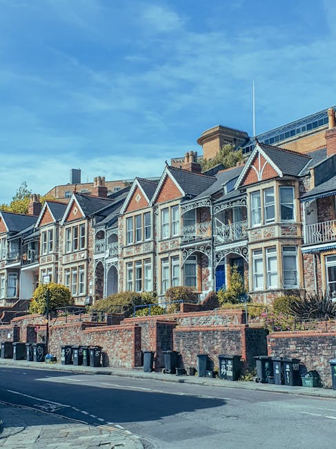 A row of Victorian-style terraced houses with ornate white metal balconies and decorative trim, set on a raised brick platform with steps leading up to the entrances. The houses feature large bay windows with white frames and tiled roofs with triangular gables. The front gardens are bordered by low brick walls, and there are small bushes and trees along the front. In the foreground, a paved street runs parallel to the houses, lined with black wheelie bins for waste collection. The scene is illuminated by natural daylight, with a bright blue sky overhead and sparse cloud cover, reflecting a quiet residential area where private waste collection or rubbish removal services like House Clearance Roehampton might operate to maintain the neighborhood's tidy appearance.