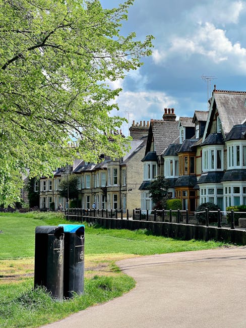 A row of Victorian-style terraced houses with ornate white metal balconies and decorative trim, set on a raised brick platform with steps leading up to the entrances. The houses feature large bay windows with white frames and tiled roofs with triangular gables. The front gardens are bordered by low brick walls, and there are small bushes and trees along the front. In the foreground, a paved street runs parallel to the houses, lined with black wheelie bins for waste collection. The scene is illuminated by natural daylight, with a bright blue sky overhead and sparse cloud cover, reflecting a quiet residential area where private waste collection or rubbish removal services like House Clearance Roehampton might operate to maintain the neighborhood's tidy appearance.